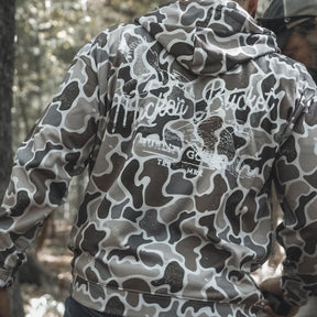 Someone wearing the Mucker Bucket Wood Ducks Premium Athletic Duck Camo Hoodie, featuring “Hacker Cracker” on the back, stands outdoors in a wooded area with their back to the camera.
