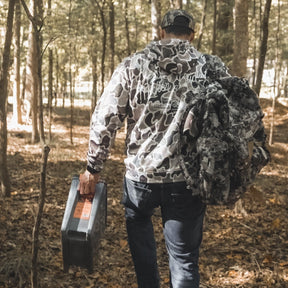 Wearing a Mucker Bucket Wood Ducks Premium Athletic Duck Camo Hoodie and cap, a person walks through a forest with a toolbox and camouflage gear, fallen leaves scattered across the ground.