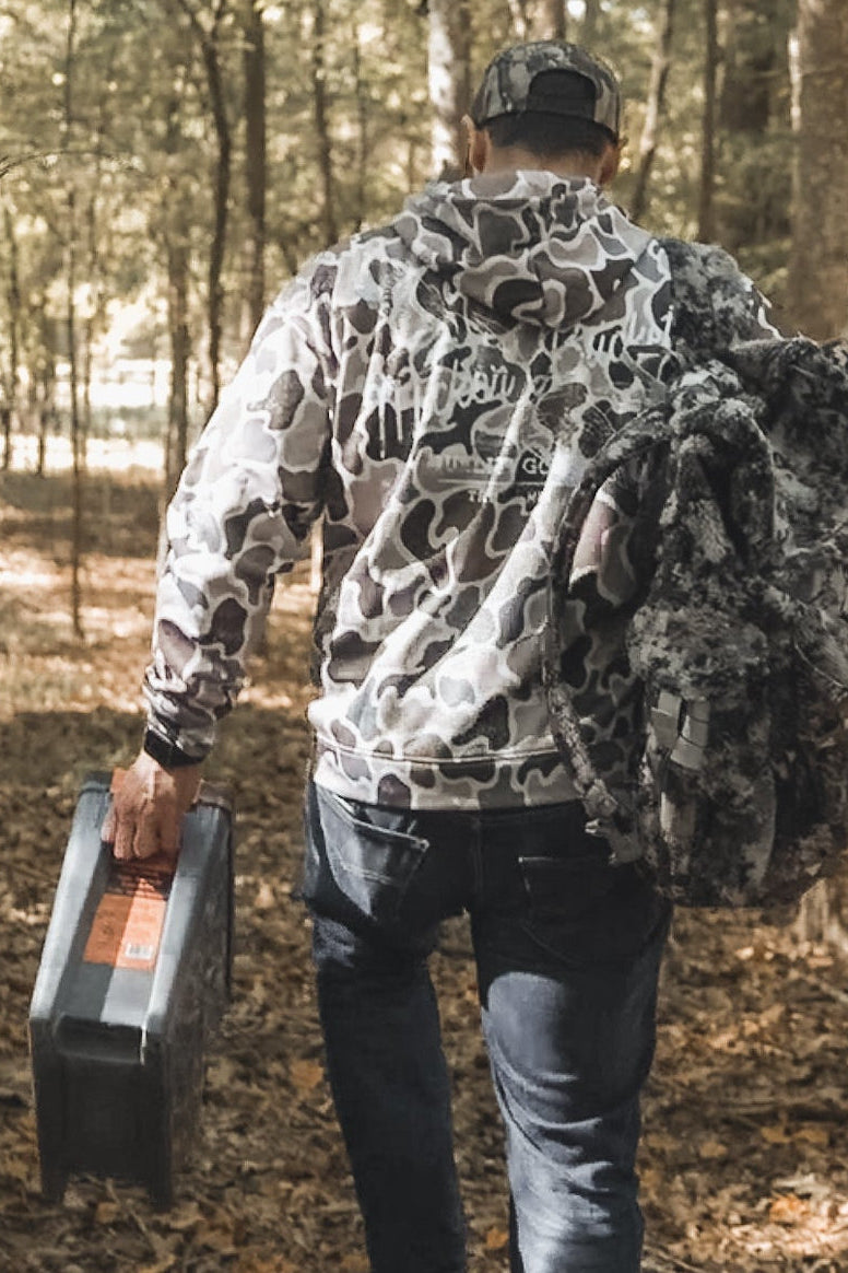 Wearing a Mucker Bucket Wood Ducks Premium Athletic Duck Camo Hoodie and cap, a person walks through a forest with a toolbox and camouflage gear, fallen leaves scattered across the ground.
