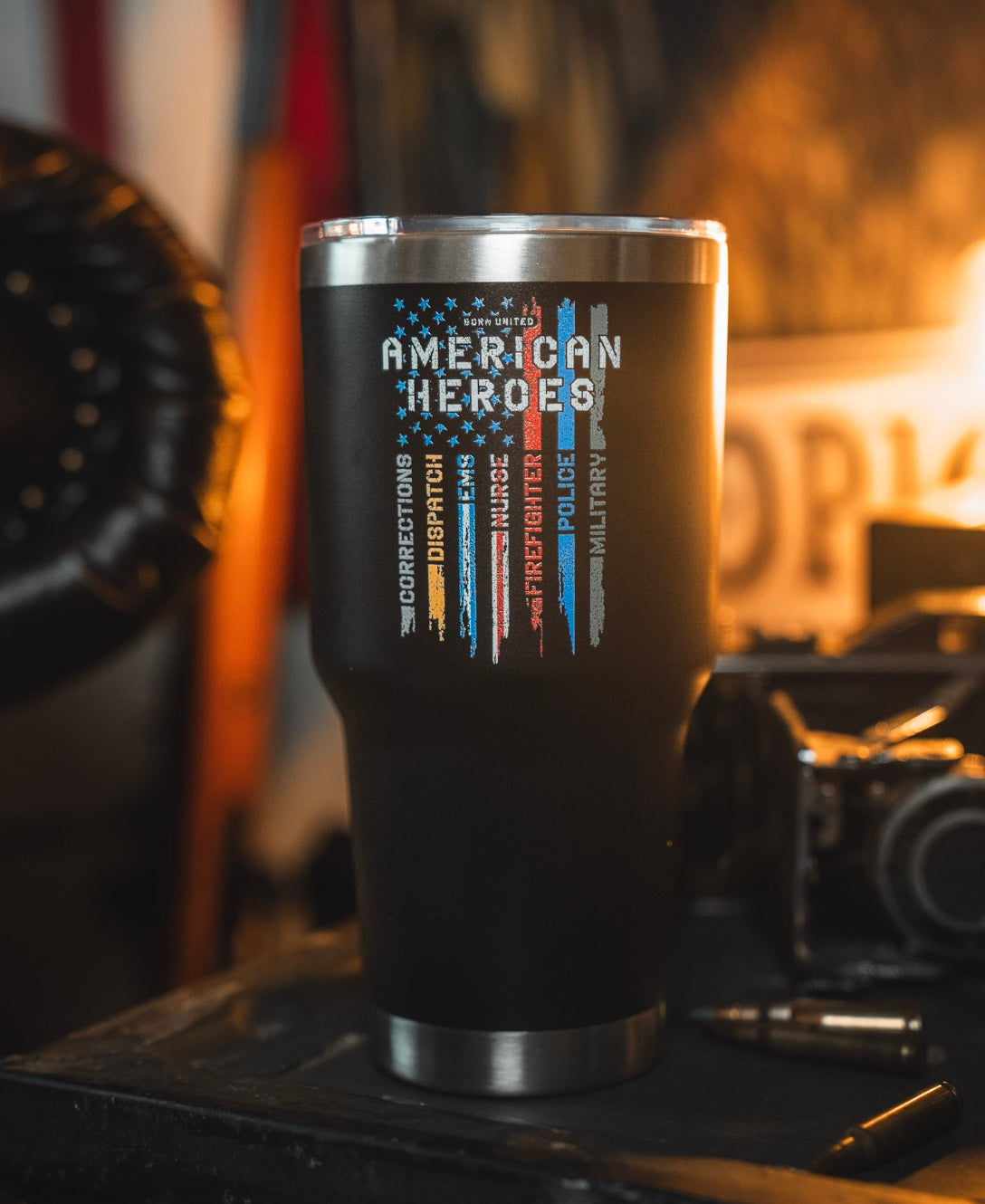 The 30oz American Heroes Tumbler by Born United Private Label, featuring colored lines for corrections, dispatch, EMS, fire, nurses, police & military, sits on a dark surface with blurred background items and a sign.