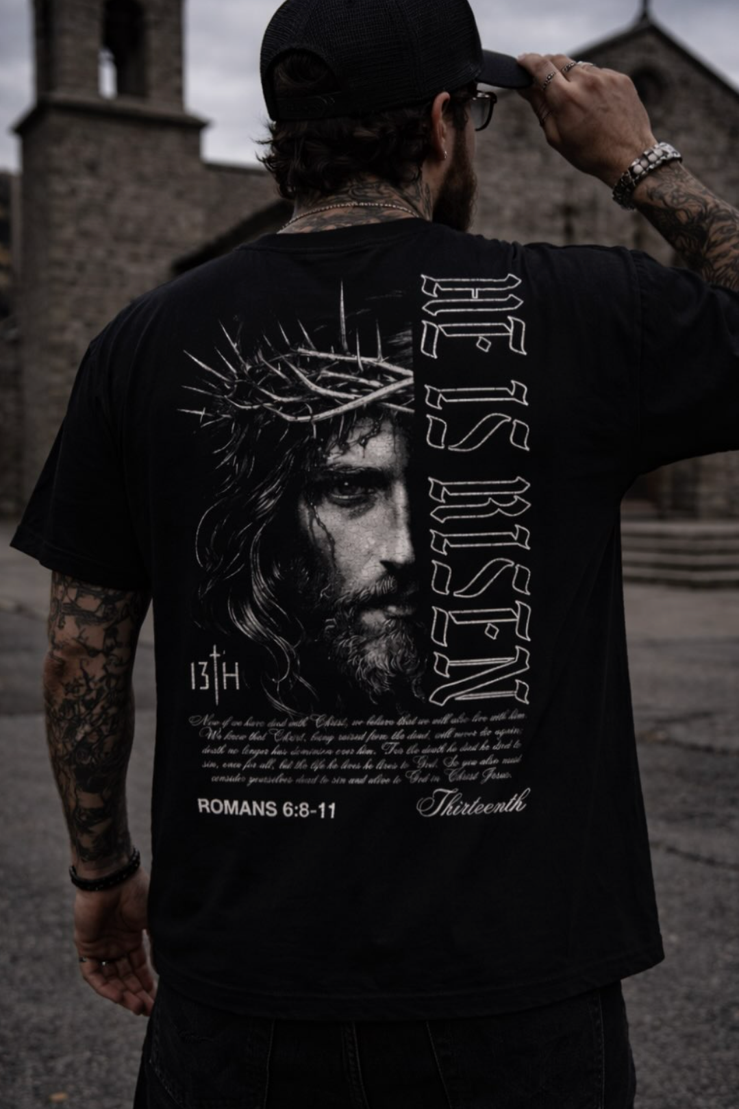 A man in a black cap and a 13th He is Risen Premium Tee, featuring an illustrated Jesus with a crown of thorns, “He is Risen” text, and a Bible verse, stands outside near stone buildings.