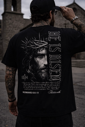 A man in a black cap and a 13th He is Risen Premium Tee, featuring an illustrated Jesus with a crown of thorns, “He is Risen” text, and a Bible verse, stands outside near stone buildings.