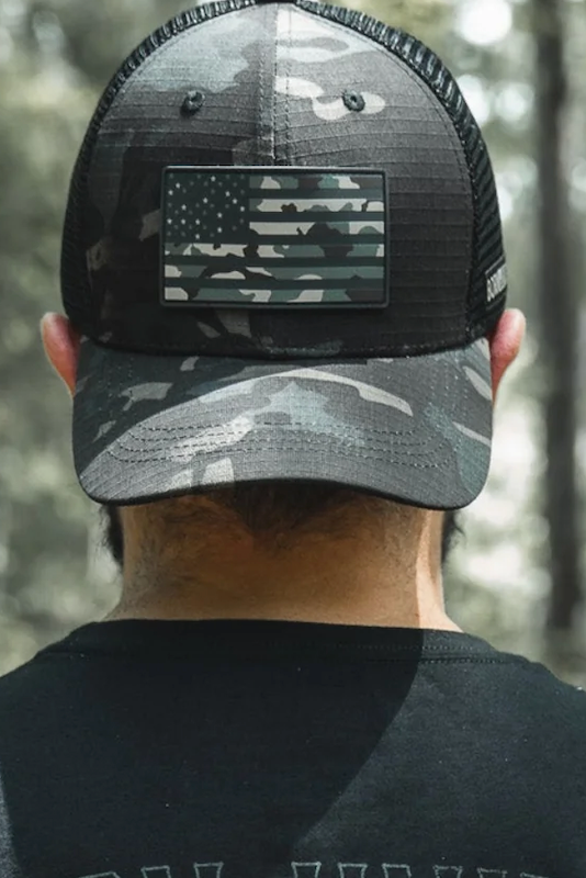 A person is seen from behind in an outdoor, wooded area wearing the Born United Private Label’s Born United Black Camo Trucker - Camo Flag cap, featuring a subdued American flag patch.