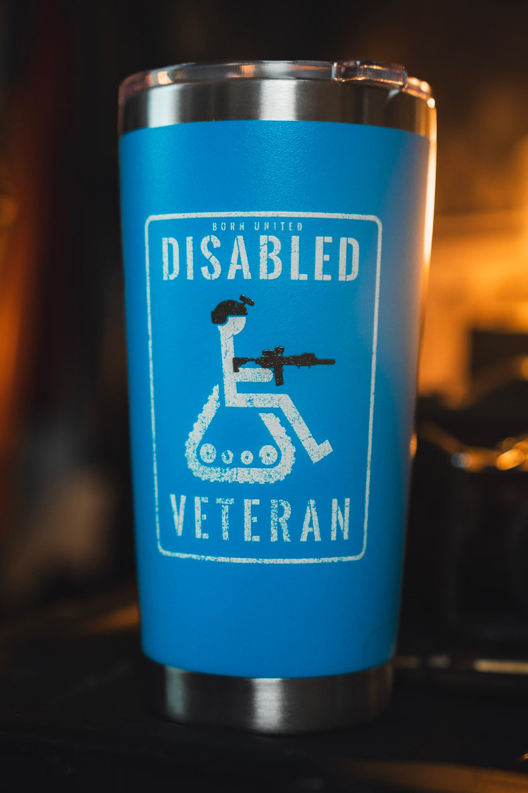 The Born United Private Label Disabled Veteran Tumbler - 20oz is blue, features a wheelchair user with a rifle and "Born United Disabled Veteran" text, has a stainless steel cupholder-friendly design, and sits on a dark surface in warm light.