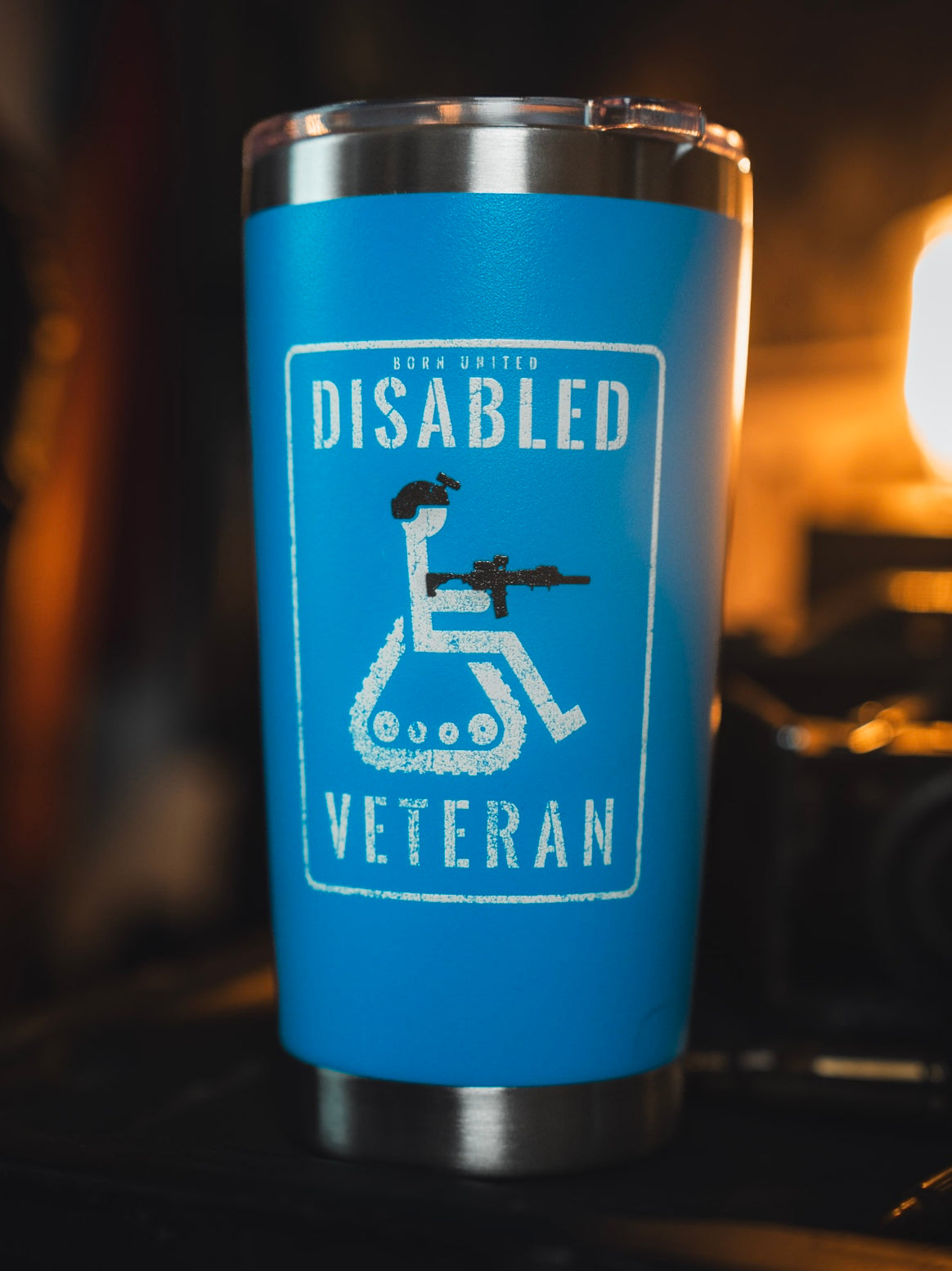 The Born United Private Label Disabled Veteran Tumbler - 20oz is blue, features a wheelchair user with a rifle and "Born United Disabled Veteran" text, has a stainless steel cupholder-friendly design, and sits on a dark surface in warm light.