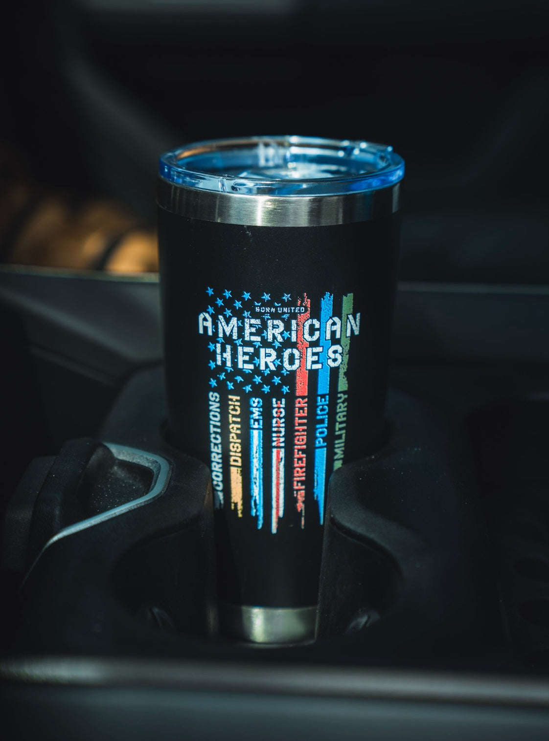 A black 20oz American Heroes Tumbler by Born United Private Label, featuring an American flag and colored stripes for police, firefighters, and military, with a blue lid, sits in a car cup holder.