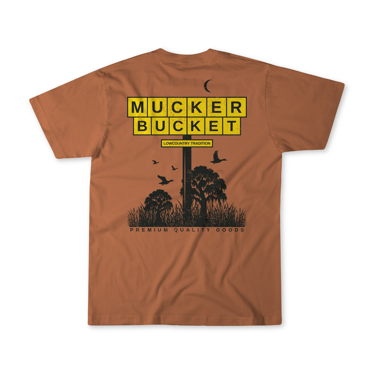 The American Tradition Premium Tee by Mucker Bucket features "MUCKER BUCKET" in bold yellow boxes, "Lowcountry Tradition" below, black tree, grass, and bird silhouettes, and "Premium Quality Goods." Made from premium ring-spun cotton for a relaxed fit.
