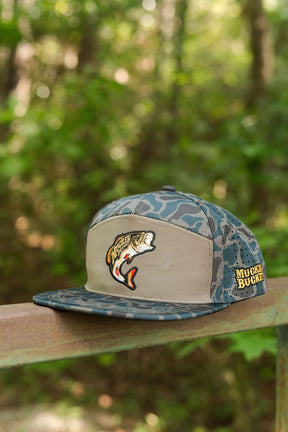 The Mucker Bucket Big Bass Daddy Hydro Duck Camo Bucket cap, featuring a fish patch and “MUCK BUCKET” embroidery, sits on a metal rail with blurred green foliage in the background.
