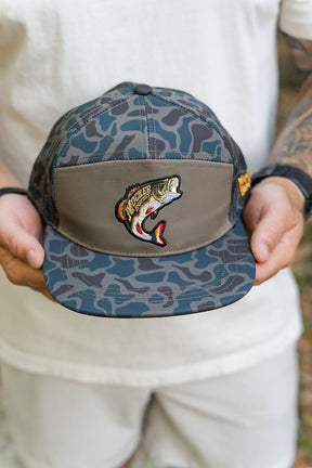 A person in a white shirt holds a gray and blue camo bucket hat labeled Big Bass Daddy Hydro Duck Camo Bucket by Mucker Bucket, featuring an embroidered fish patch on the front.