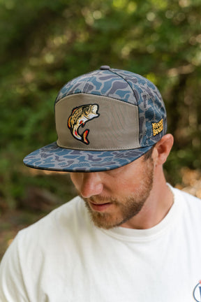 A bearded man wears a high-quality blue and gray camo-patterned Big Bass Daddy Hydro Duck Camo Bucket by Mucker Bucket, featuring an embroidered fish logo patch with Mucho Bueno, standing outdoors with greenery in the background.
