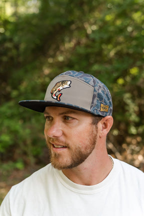 A bearded man in a white shirt stands outdoors, wearing the Mucker Bucket Big Bass Daddy Hydro Duck Camo Bucket hat featuring a fish logo and an embroidered Muck Bucket patch, set against a blurred green leafy background.