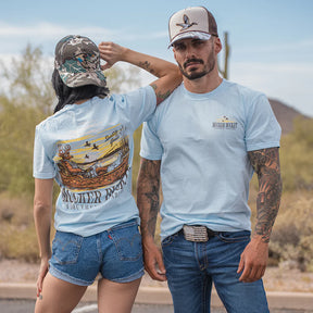 A woman and man outdoors wear Mucker Bucket Buck Dreamin' Premium Tees in light blue; she faces away to display the graphic on the back, while he faces forward showing the small logo on his premium cotton t-shirt.