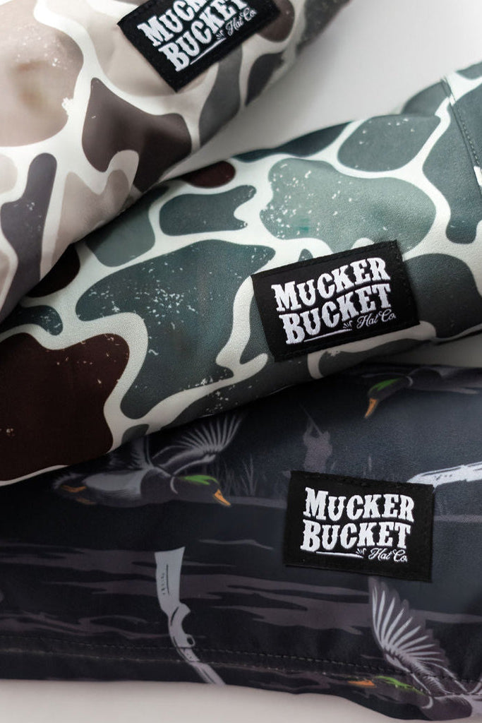 Three rolled-up garments with camouflage and duck patterns, labeled Mucker Bucket on a black tag, in shades of brown, green, and gray are from the Mens Performance Athletic Shorts line titled Duck, Duck Boom.