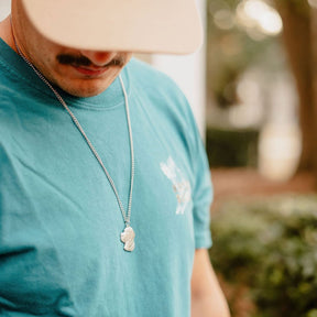 A person in a Mucker Bucket Wood Ducks Premium Blue Tee, beige cap, and silver dog-shaped pendant necklace looks down amid greenery and soft outdoor light.