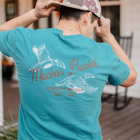 A person in the Mucker Bucket Wood Ducks Premium Blue Tee, featuring illustrated ducks and branding on the back, adjusts their cap while standing on a porch.