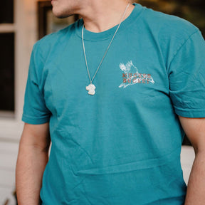A person wears the Mucker Bucket Wood Ducks Premium Blue Tee, crafted from premium cotton in blue with a graphic chest design, paired with a silver bear-shaped pendant necklace. The garment dyed shirt stands out against a blurred background.