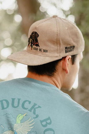 A man wears the Mucker Bucket Good Ol' Boy Tan Corduroy snapback hat with embroidered text and a dog image on the back, paired with a light blue-green shirt featuring a flying duck and “DUCK, BO,” creating a relaxed outdoor vibe.