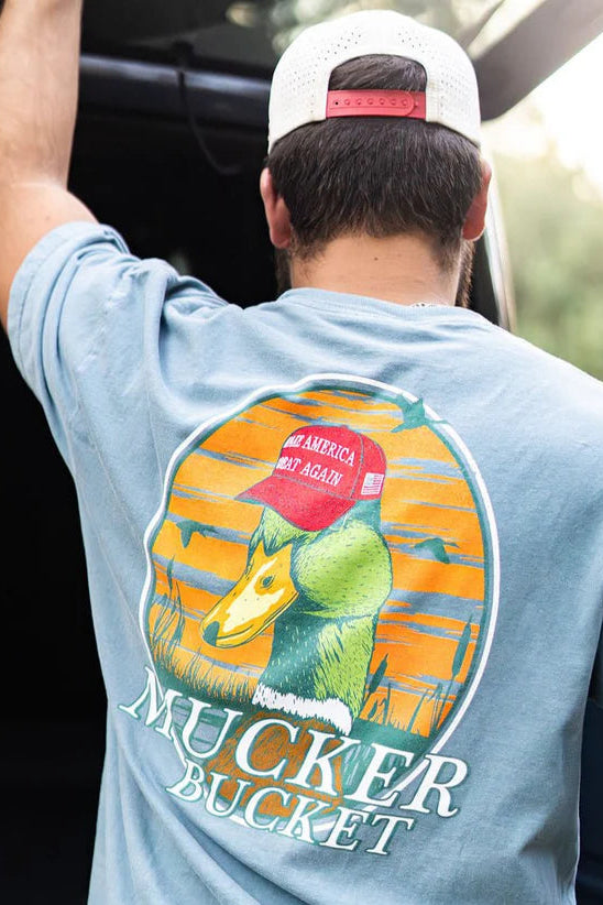 A man in a cap and light blue Smart Duck Tee by Mucker Bucket with a duck graphic wearing a red hat stands against blurred greenery and part of a car. The ring-spun cotton shirt offers a premium relaxed fit.