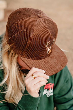 A person in the Mucker Bucket Monster Buck Corduroy Bucket hat with an embroidered antler design tilts their head down, partially hiding their face. Their long blond hair is visible, and they wear a green hoodie with a red and white logo.