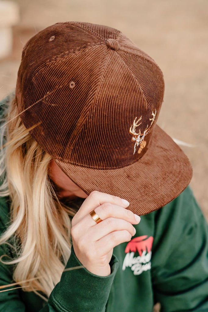 A person in the Mucker Bucket Monster Buck Corduroy Bucket hat with an embroidered antler design tilts their head down, partially hiding their face. Their long blond hair is visible, and they wear a green hoodie with a red and white logo.