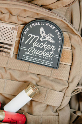 Tan tactical backpack with American flag patch and 'Mucker Bucket' logo, featuring shotgun shells and a knife.
