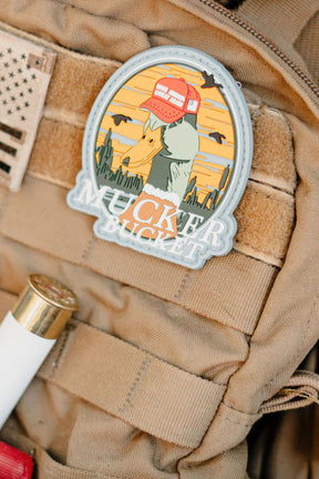 The Mucker Bucket Smart Duck PVC Patch, a 4x4 Velcro patch with a dog in a red cap against a sunset desert, is attached to a tan tactical vest beside a shotgun shell and an American flag patch.