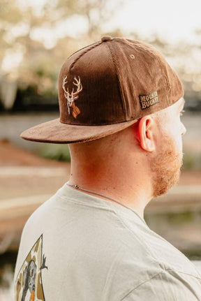 A man with a beard stands outdoors near water, facing away from the camera, wearing the Mucker Bucket Monster Buck Corduroy Bucket hat.