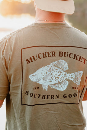 Man wearing a 'Mucker Bucket' t-shirt with a fish graphic outdoors.