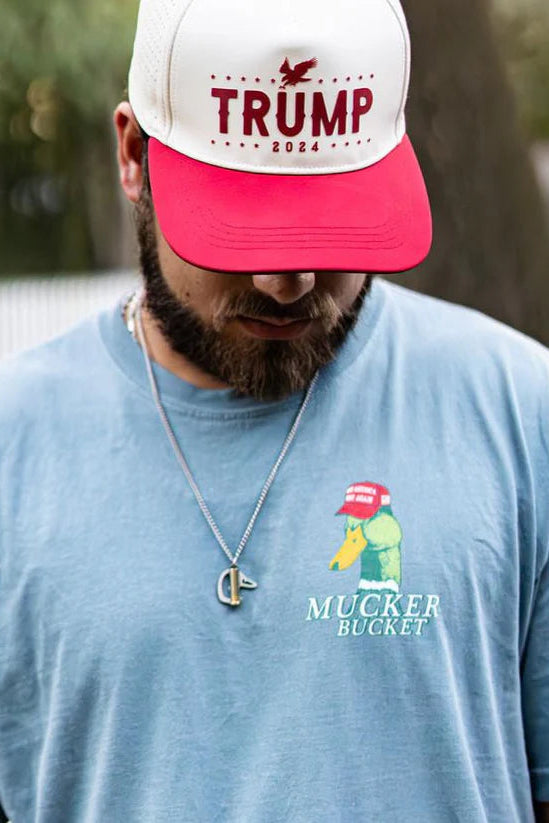 Wearing a light blue premium relaxed fit Smart Duck Tee by Mucker Bucket, featuring a cartoon duck with a red cap, a man glances down. His red and white cap reads Trump 2024, while a chain necklace rests on the garment-dyed 100% ring-spun cotton fabric.