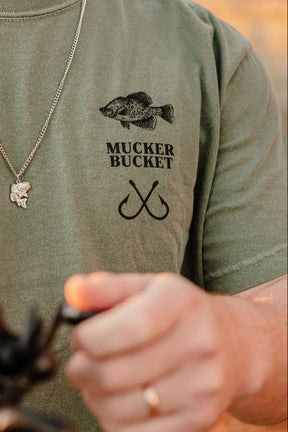 A person wears the Crappie Premium Tee by Mucker Bucket in olive-green, featuring a fish graphic, "MUCKER BUCKET" text, and two fishing hooks, accessorized with a fish-shaped necklace and gold ring. Their hand is shown in the foreground.