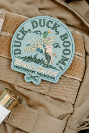 The Duck Duck Boom PVC Patch by Mucker Bucket features flying ducks, the phrase “Duck Duck Boom!”, and a shotgun shell on tan fabric, capturing a classic lowcountry hunting tradition.