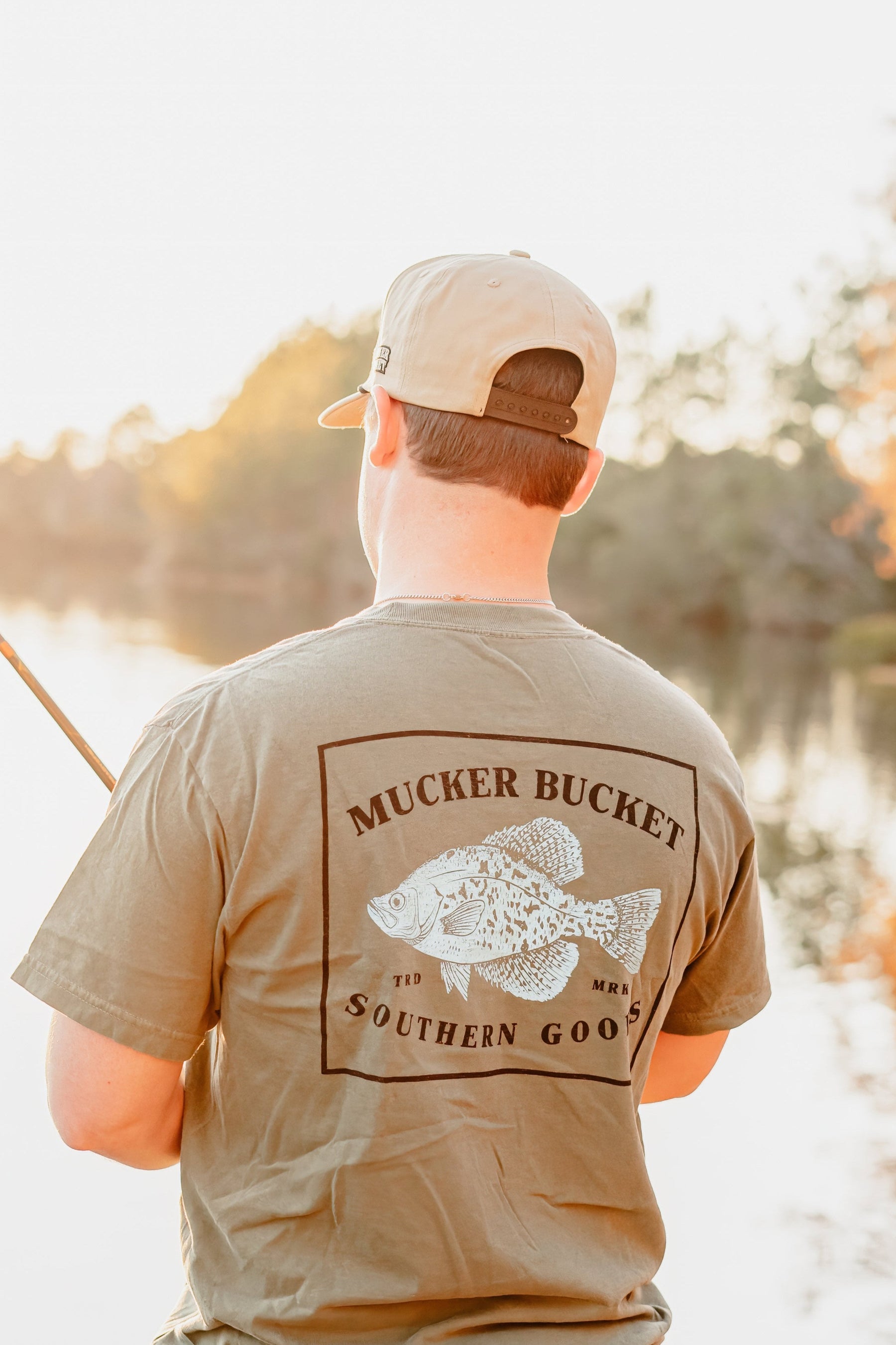 A person in a khaki cap and Mucker Bucket Crappie Premium Tee with a fish graphic is fishing by a calm lake at sunset, surrounded by soft sunlight and trees.