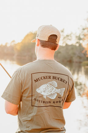 A person in a khaki cap and Mucker Bucket Crappie Premium Tee with a fish graphic is fishing by a calm lake at sunset, surrounded by soft sunlight and trees.