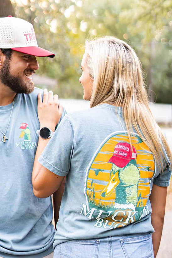 A smiling couple outdoors wears matching Mucker Bucket Smart Duck Tees, crafted from 100% ring-spun cotton with colorful back graphics. The man dons a red and white cap, while the woman displays her Apple Watch amid trees and a house in a sunny, relaxed setting.