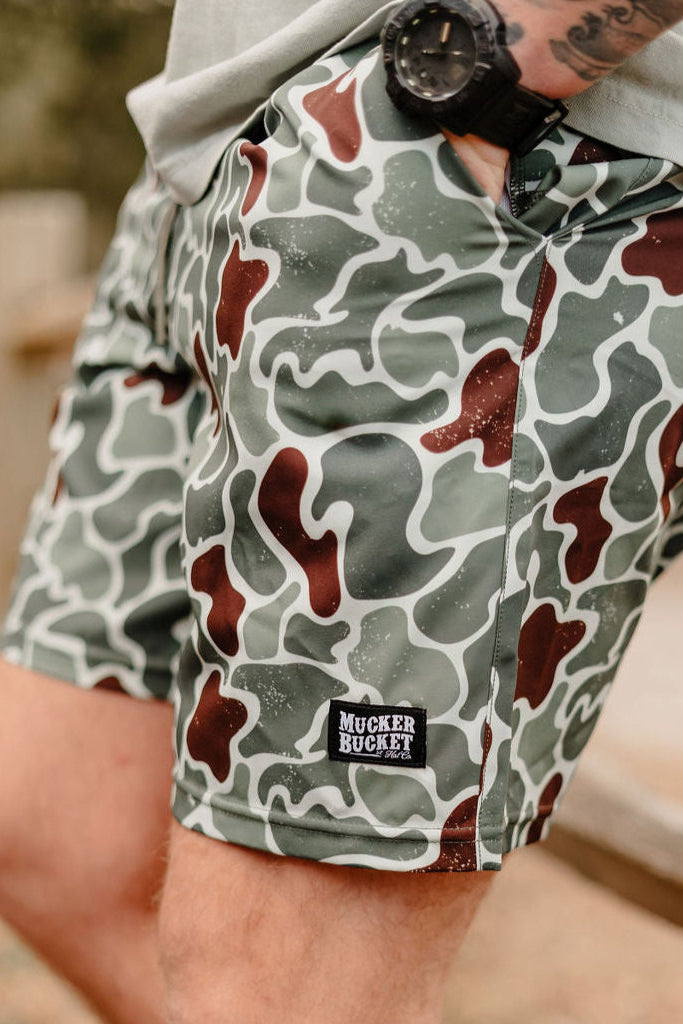 A person wearing Mucker Buckets Mens Performance Athletic Shorts, 5.5- Duck King in green and brown camo-pattern, a watch, and a light shirt is leaning on a wooden surface outdoors.