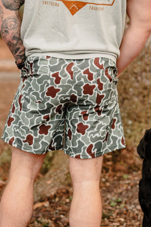 A person wearing a light gray shirt and Mucker Buckets Mens Performance Athletic Shorts in Duck King camo stands next to a black dog. The tattooed individual is outdoors by a tree, as the dog looks toward the camera.