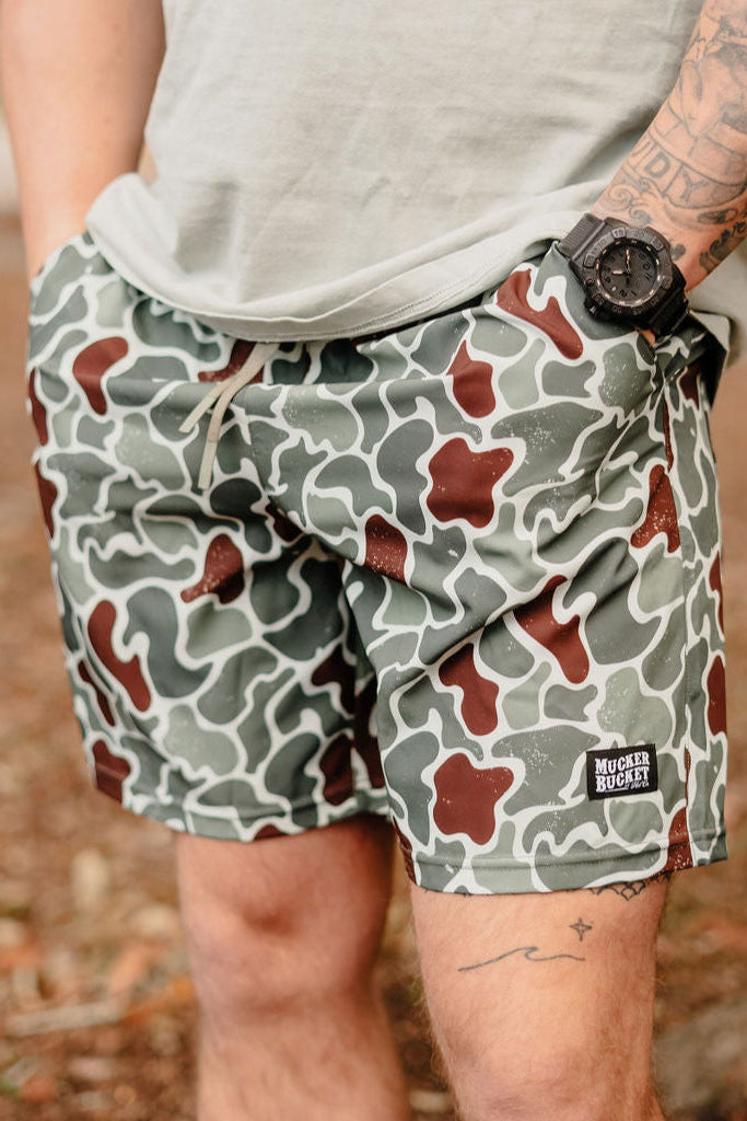 A person wearing Mens Performance Athletic Shorts | 5.5- Duck King by Mucker Bucket has hands in pockets, with a watch and tattoos visible on their left arm against a backdrop of blurred foliage and ground.