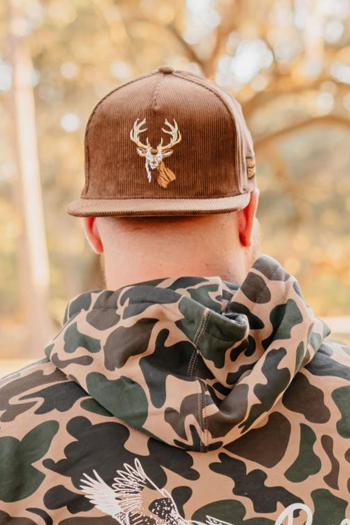 A person seen from behind wears the Mucker Bucket Monster Buck Corduroy Bucket hat, featuring embroidered deer antlers, and a camouflage hoodie with a bird graphic. The stylish hat stands out against the softly blurred outdoor background.