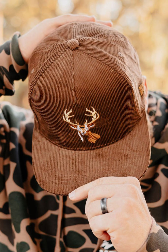 Someone wearing a Mucker Bucket Monster Buck Corduroy Bucket hat with deer antler and leaf pins, paired with a camouflage hoodie, is holding the flat brim of the cap.