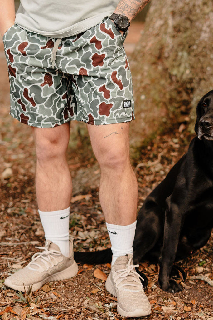 A person in Mucker Bucket Mens Performance Athletic Shorts | 5.5- Duck King stands beside a black Labrador on the ground, wearing beige sneakers and white socks, with trees and fallen leaves in the background.