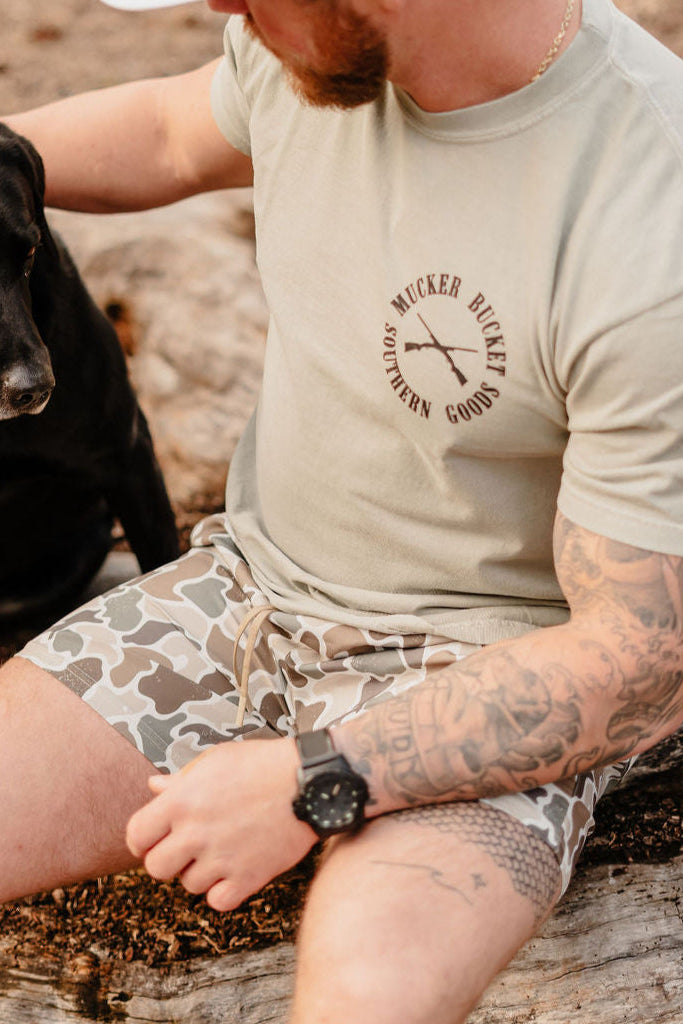 A tattooed man in a beige T-shirt and Mucker Buckets Duck It shorts sits on a log with his calm black Labrador in an outdoor setting, both seemingly savoring the peaceful moment.
