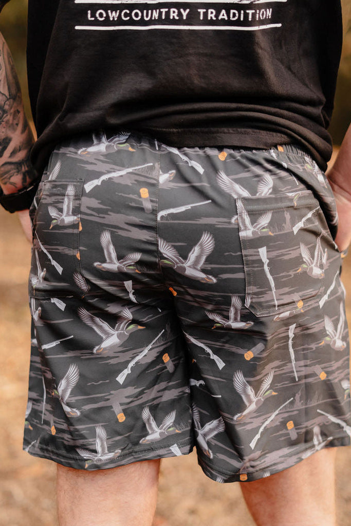 A person stands outdoors in a black t-shirt with LOWCOUNTRY TRADITION and Mucker Buckets Mens Performance Athletic Shorts | 5.5- Duck, Duck Boom, featuring flying bird designs and orange dots. They have tattoos on their left arm and hands in pockets against a blurred natural background.