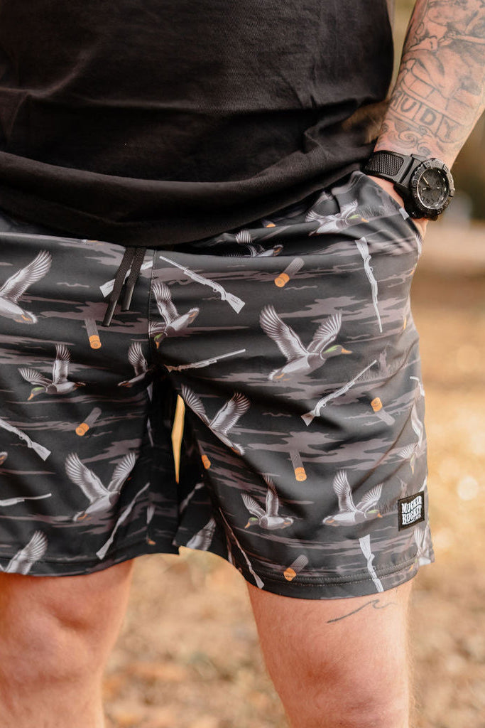 A person outdoors in Mucker Buckets Mens Performance Athletic Shorts, 5.5- Duck, Duck Boom, with a crane pattern and dark drawstring, wears a black watch and T-shirt. The earthy-toned background is blurred.