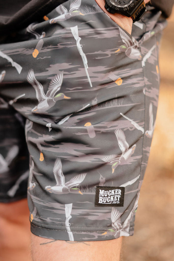 A person outdoors is wearing Mucker Buckets Mens Performance Athletic Shorts in the Duck, Duck Boom pattern, featuring gray camouflage with ducks and shotguns. Part of their hand is visible.