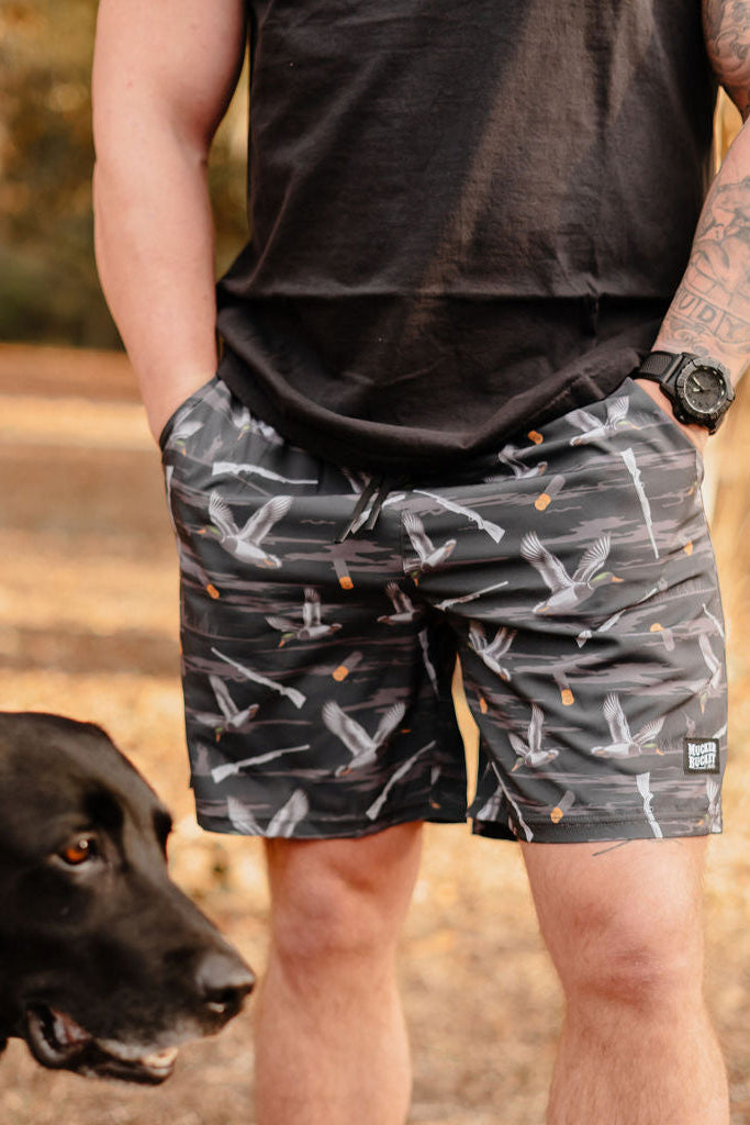 A person in a black shirt and Mucker Buckets Mens Performance Athletic Shorts | 5.5- Duck, Duck Boom stands outdoors with hands in pockets. A black dog is beside them, against a backdrop of trees and soft sunlight.