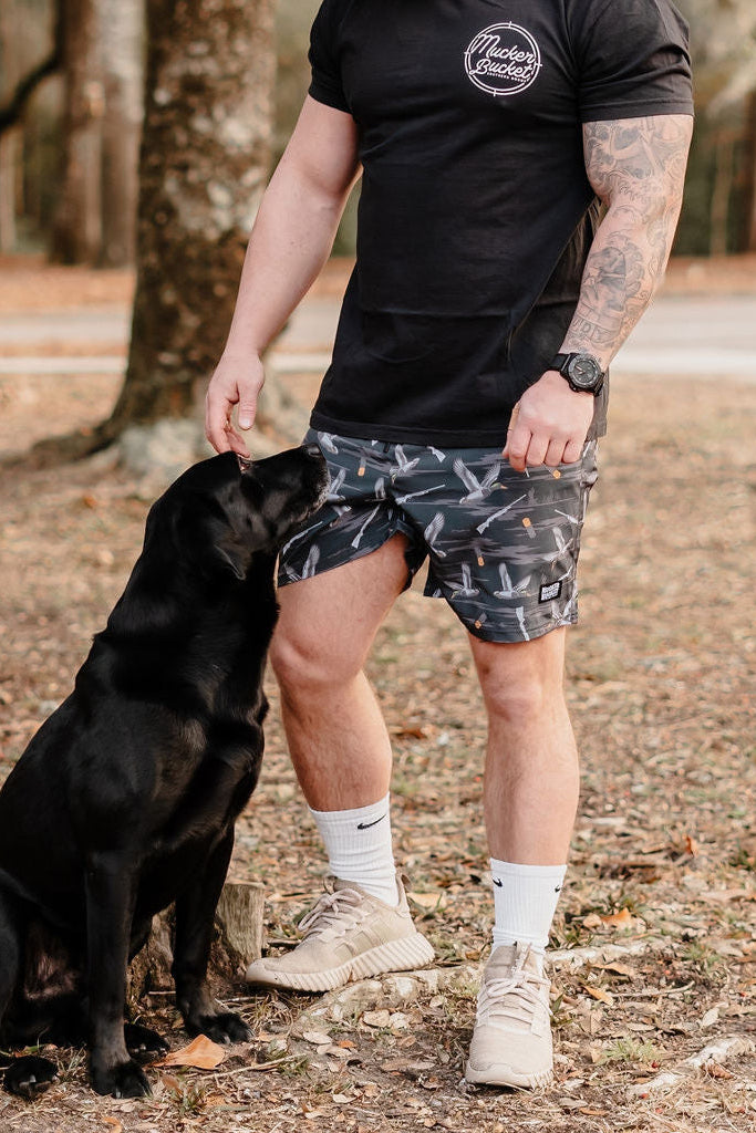 A man in a black t-shirt and Mucker Buckets Mens Performance Athletic Shorts | 5.5- Duck, Duck Boom stands outdoors petting a black Labrador beside him, surrounded by trees and scattered dry leaves.