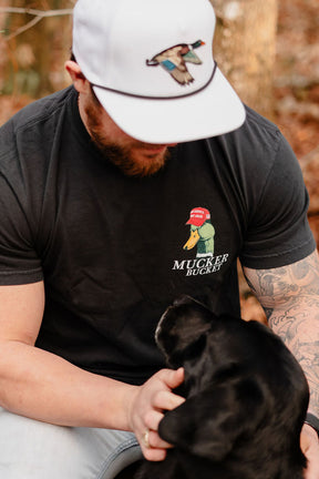Wearing a Mucker Bucket relaxed fit Smart Duck - Black tee in soft ring-spun cotton, a man sits outdoors in a white hat with a bird logo, looking down and petting a black dog. Brown-leaved trees are visible behind him.