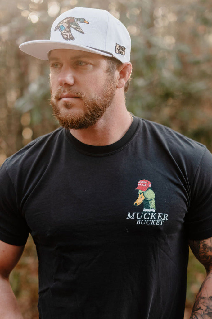 A bearded man stands outdoors in a white hat and the Mucker Bucket Smart Duck - Black relaxed fit tee, featuring a duck with a red cap and "MUCKER BUCKET" text, against a backdrop of blurred greenery.