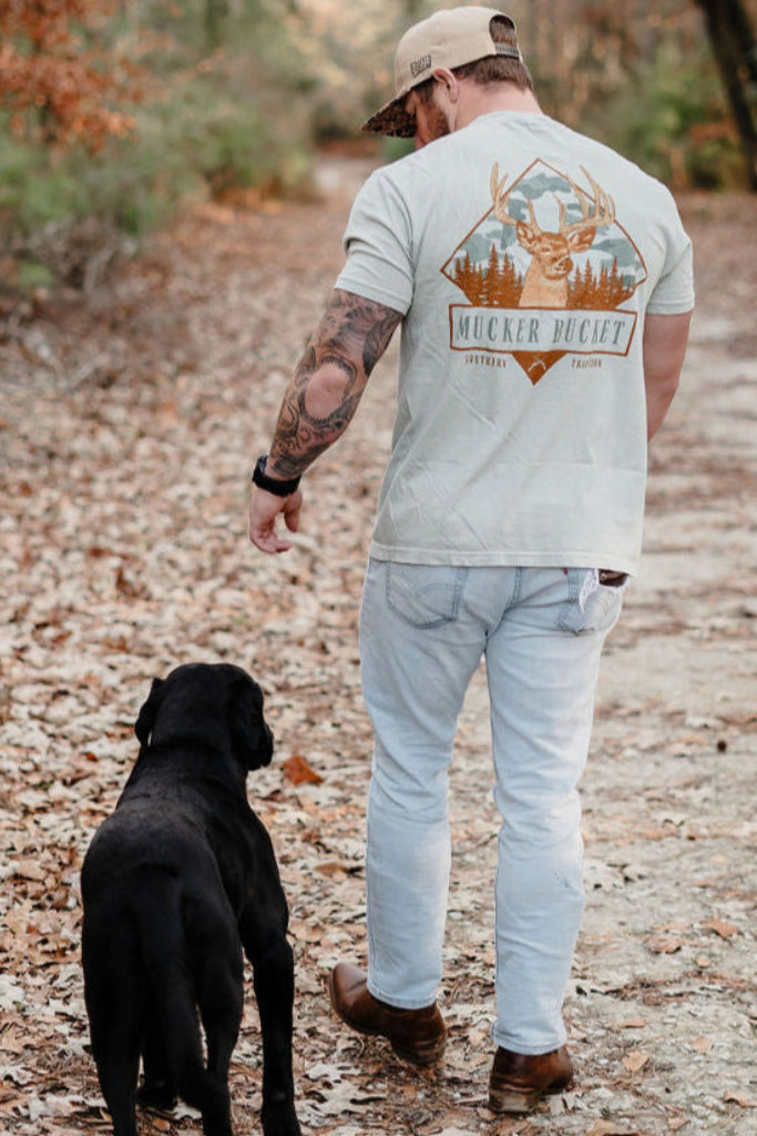 A man in jeans and a cap walks a black dog down a leafy path, wearing the Buck Season Premium Tee by Mucker Bucket, featuring a White Tail buck graphic with antlers, set in an outdoor wooded area.