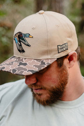 A bearded man looks down, wearing the Mucker Bucket Mallard Old School Duck Bucket hat featuring a flying mallard embroidery on camo; "Mucker Bucket" is stitched on the side—ideal for hunting season.